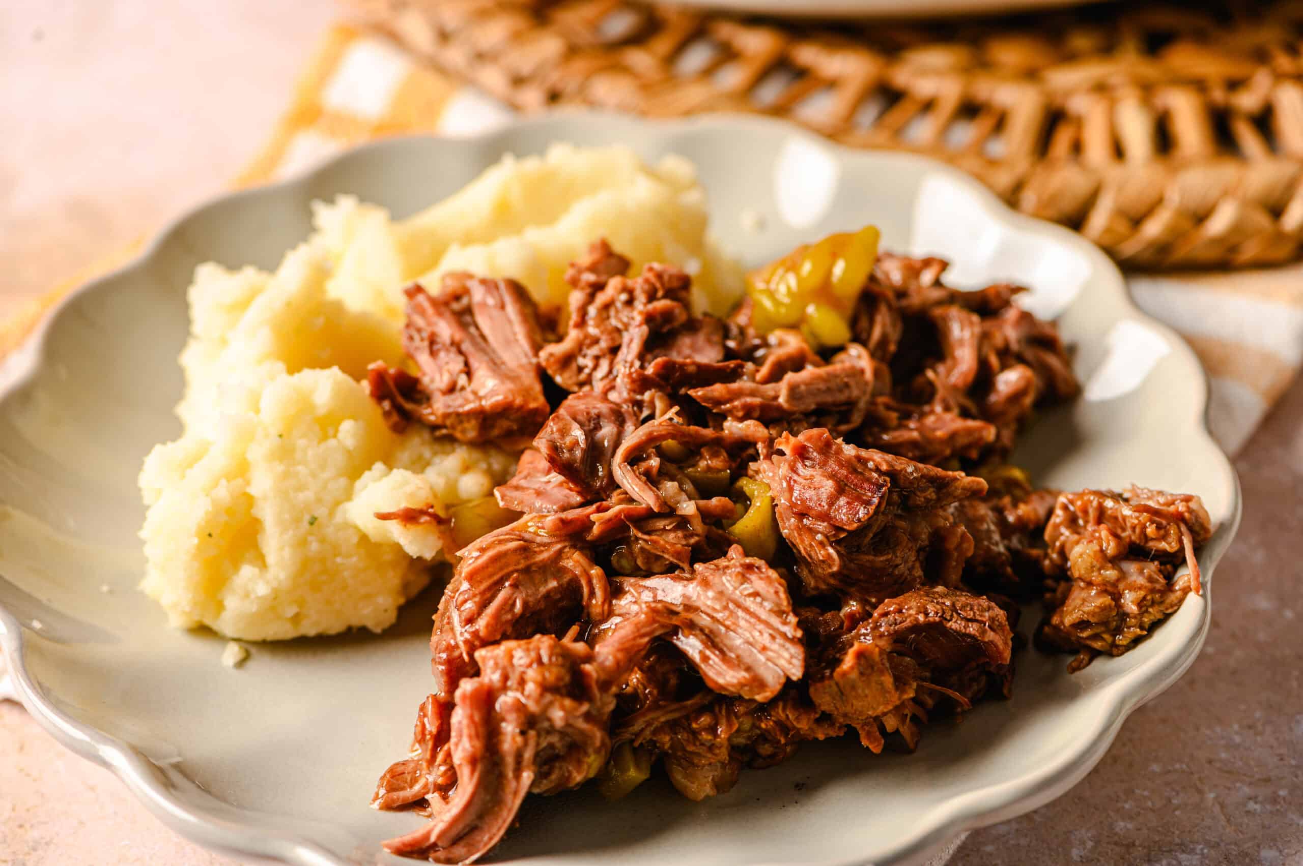 Slow cooker shredded beef with mashed potatoes on a white decorative plate, served with green pickled peppers, ready for a hearty meal.