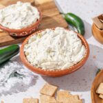 Cream cheese spread with herbs in terracotta bowl on white speckled countertop, accompanied by crackers, jalapeño, and wooden bowls of snacks, ready for serving.