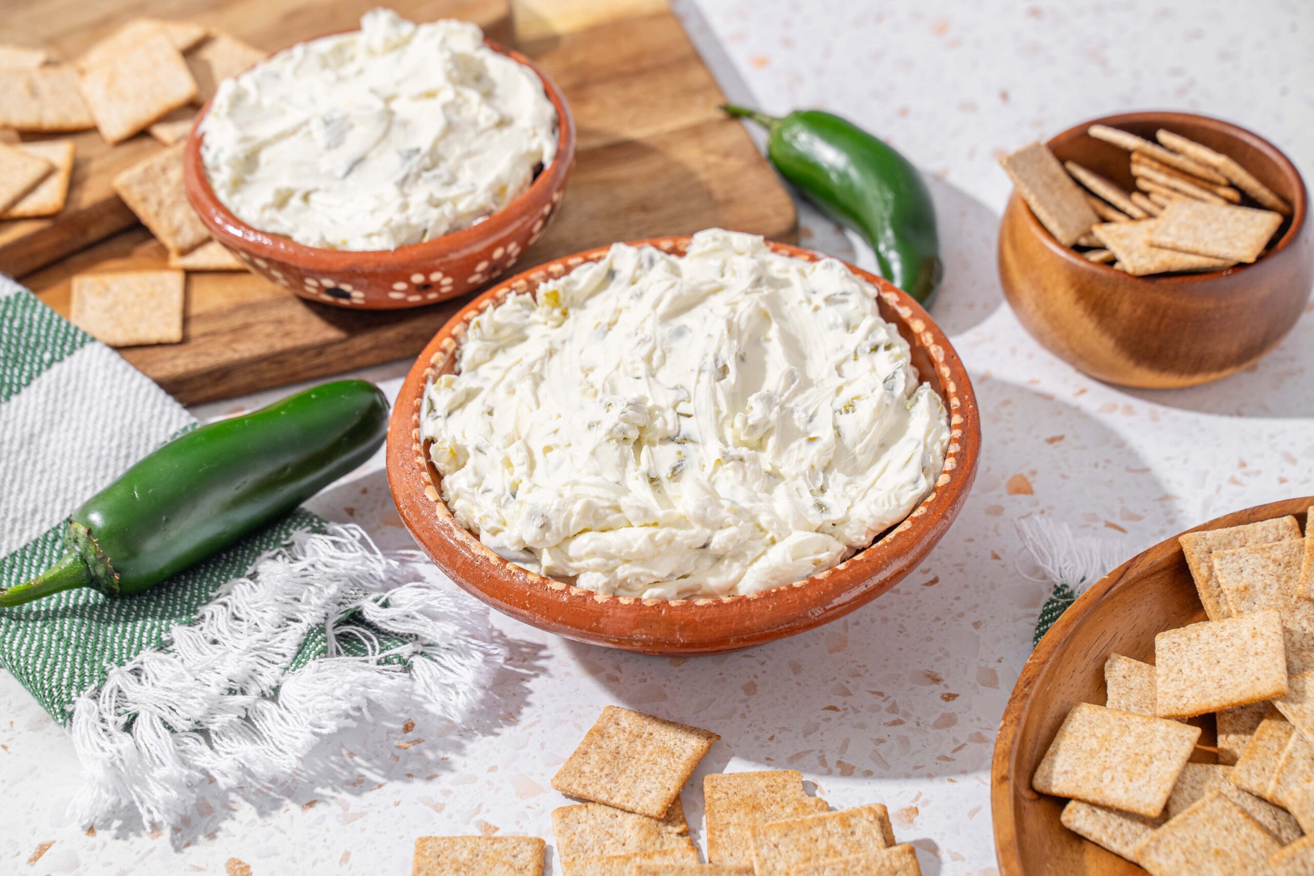 Cream cheese spread with herbs in terracotta bowl on white speckled countertop, accompanied by crackers, jalapeño, and wooden bowls of snacks, ready for serving.