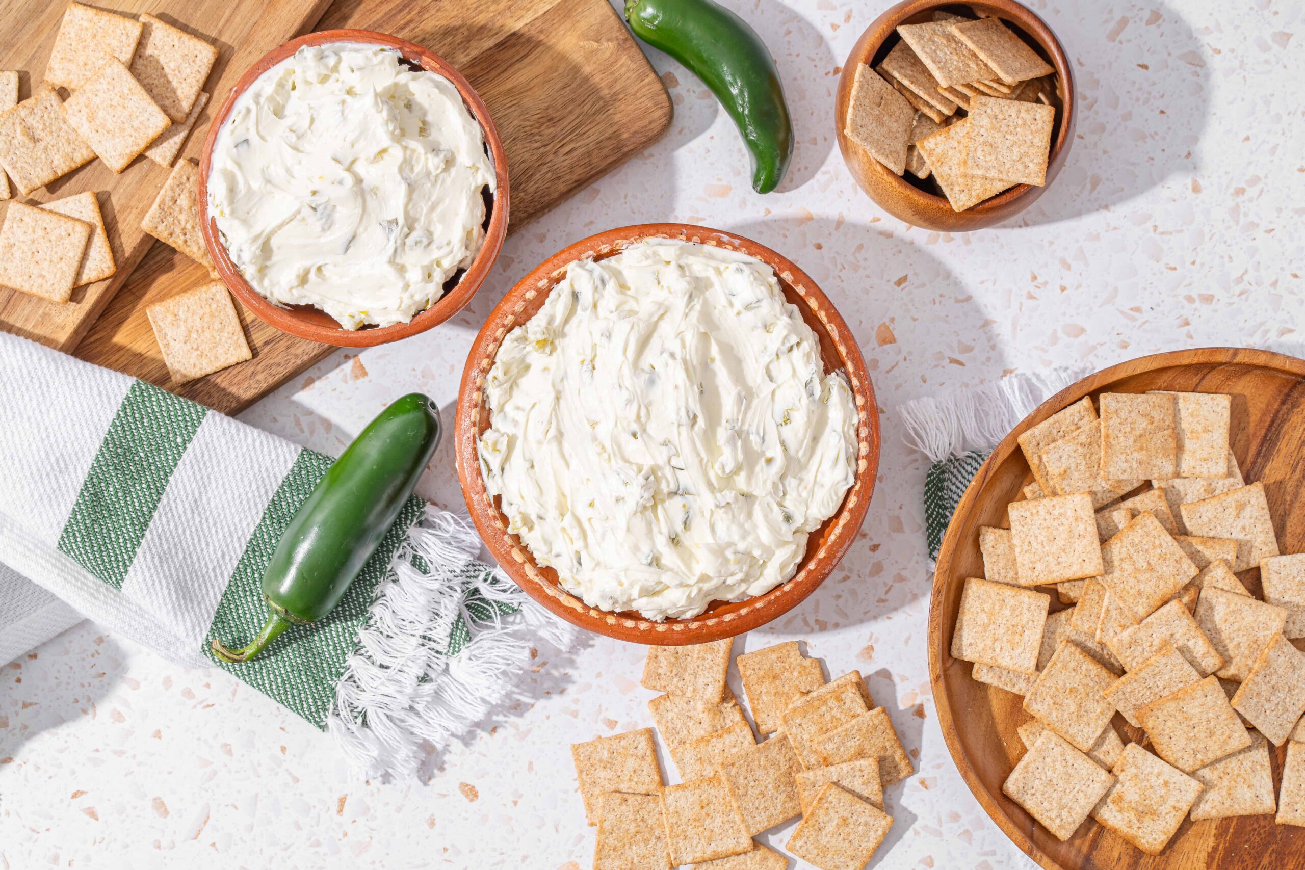 Cream cheese dip with crispy crackers and fresh jalapeño peppers on a white textured surface, featuring wooden bowls, a kitchen towel, and a cutting board for a delicious appetizer setup.