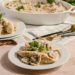 Creamy chicken and mushroom casserole served on a white plate with a sprig of parsley, with additional casserole and ingredients in the background on a rustic table setting.