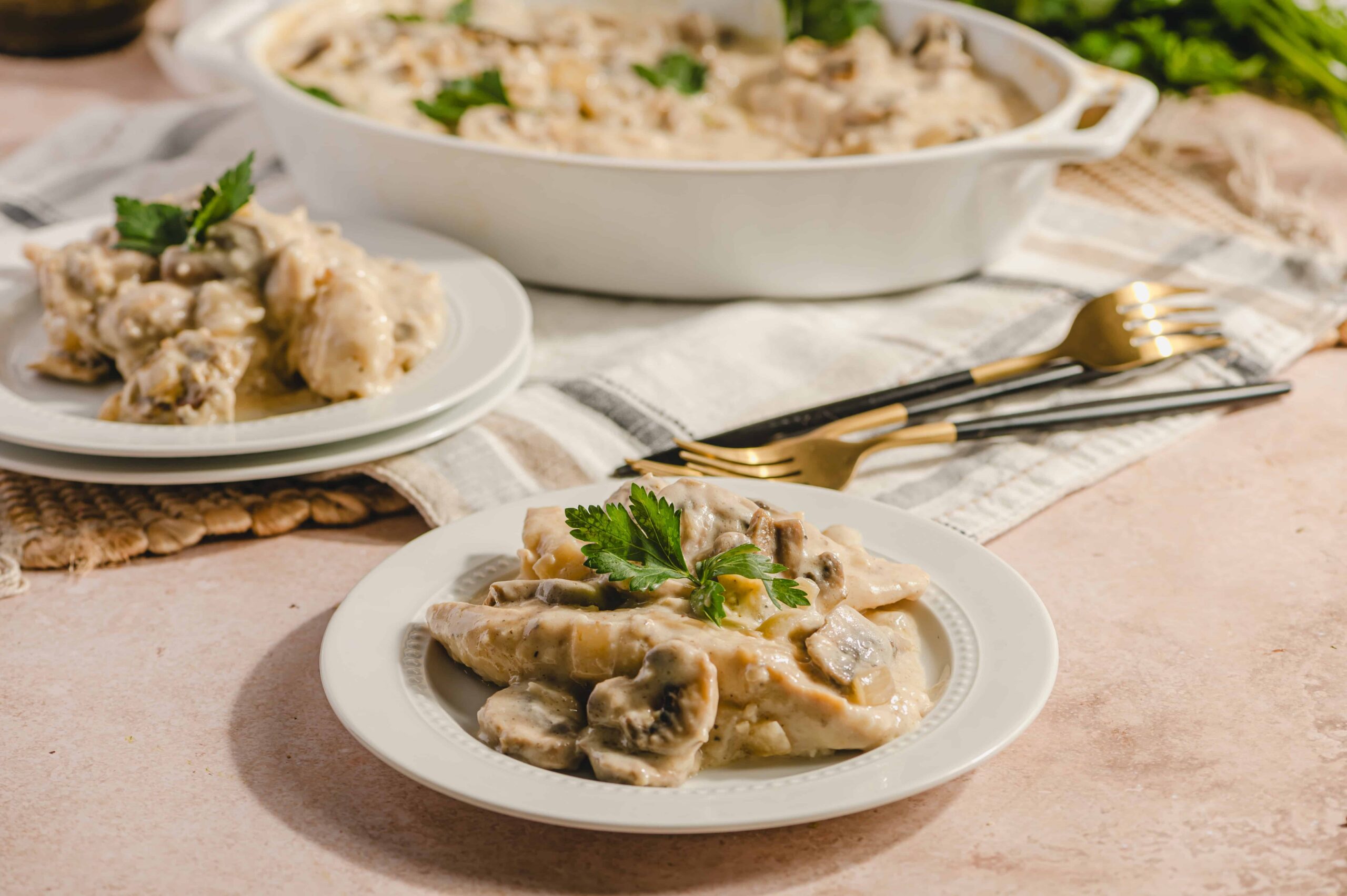 Creamy chicken and mushroom casserole served on a white plate with a sprig of parsley, with additional casserole and ingredients in the background on a rustic table setting.