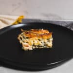 Cheesy vegetable lasagna slice on a black plate with a marble background and fabric napkin for a cozy, home-cooked meal presentation.