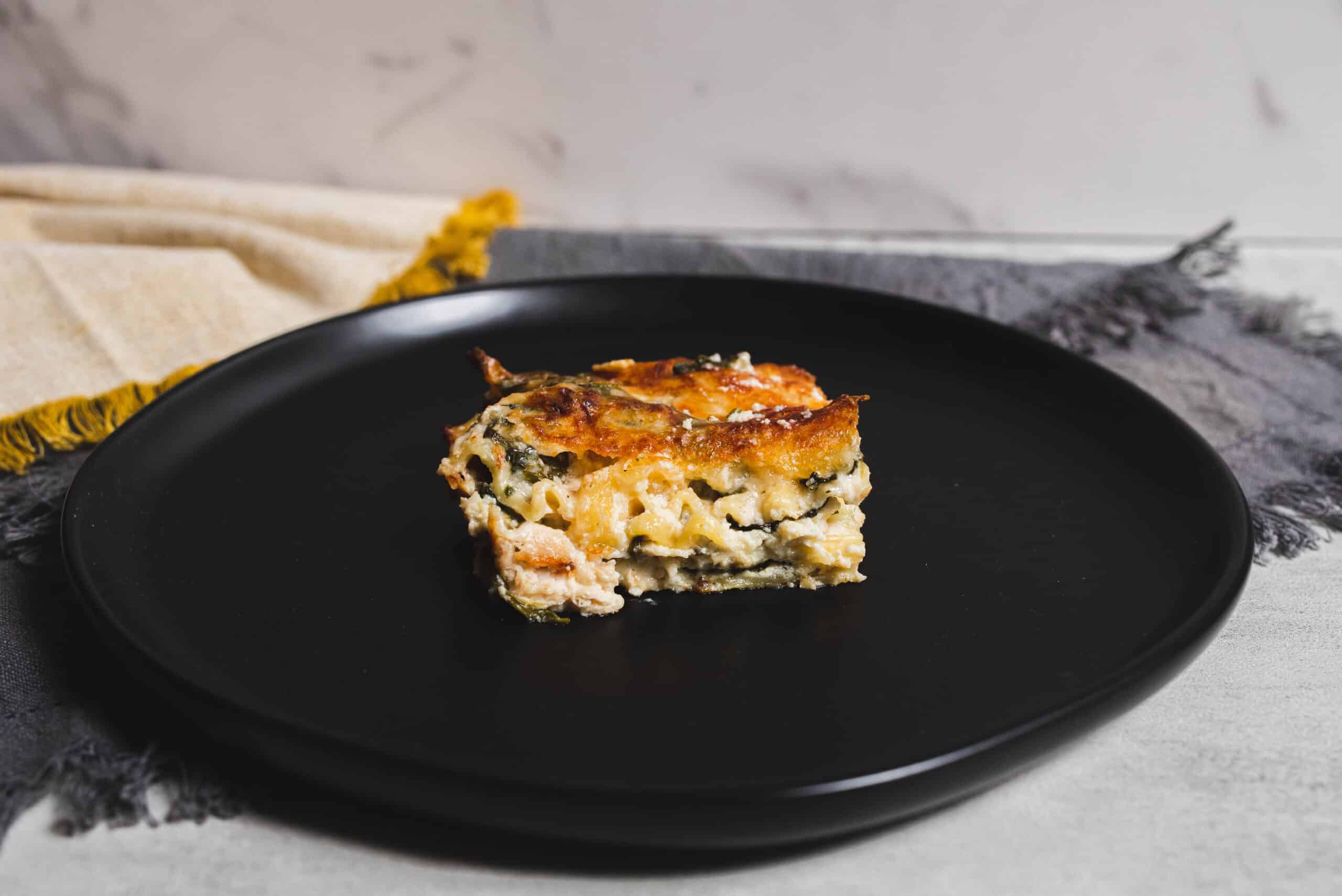 Cheesy vegetable lasagna slice on a black plate with a marble background and fabric napkin for a cozy, home-cooked meal presentation.