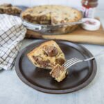Chocolate chip cookie dough cheesecake slice on a dark plate with a fork, featuring gooey chocolate chunks and creamy layers, served with a whole cheesecake in the background.