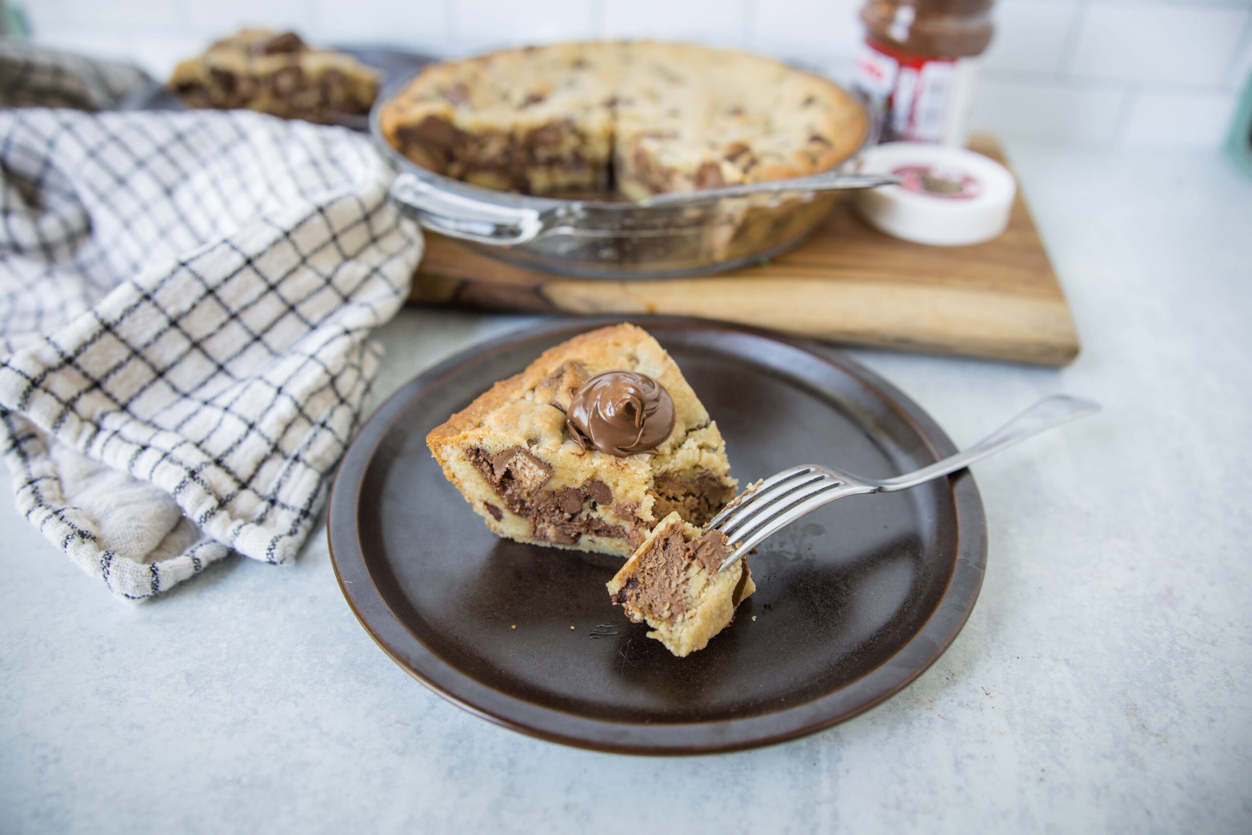 Chocolate chip cookie dough cheesecake slice on a dark plate with a fork, featuring gooey chocolate chunks and creamy layers, served with a whole cheesecake in the background.