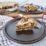 Cream cheese and chocolate chip cookie dough cheesecake topped with Nutella on a dark plate.