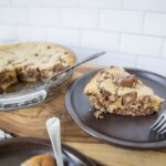 Chunks of homemade chocolate chip cookie dough cheesecake on a dark plate with a fork, with the full pie in a glass baking dish in the background, on a rustic wooden table with a white tiled wall.