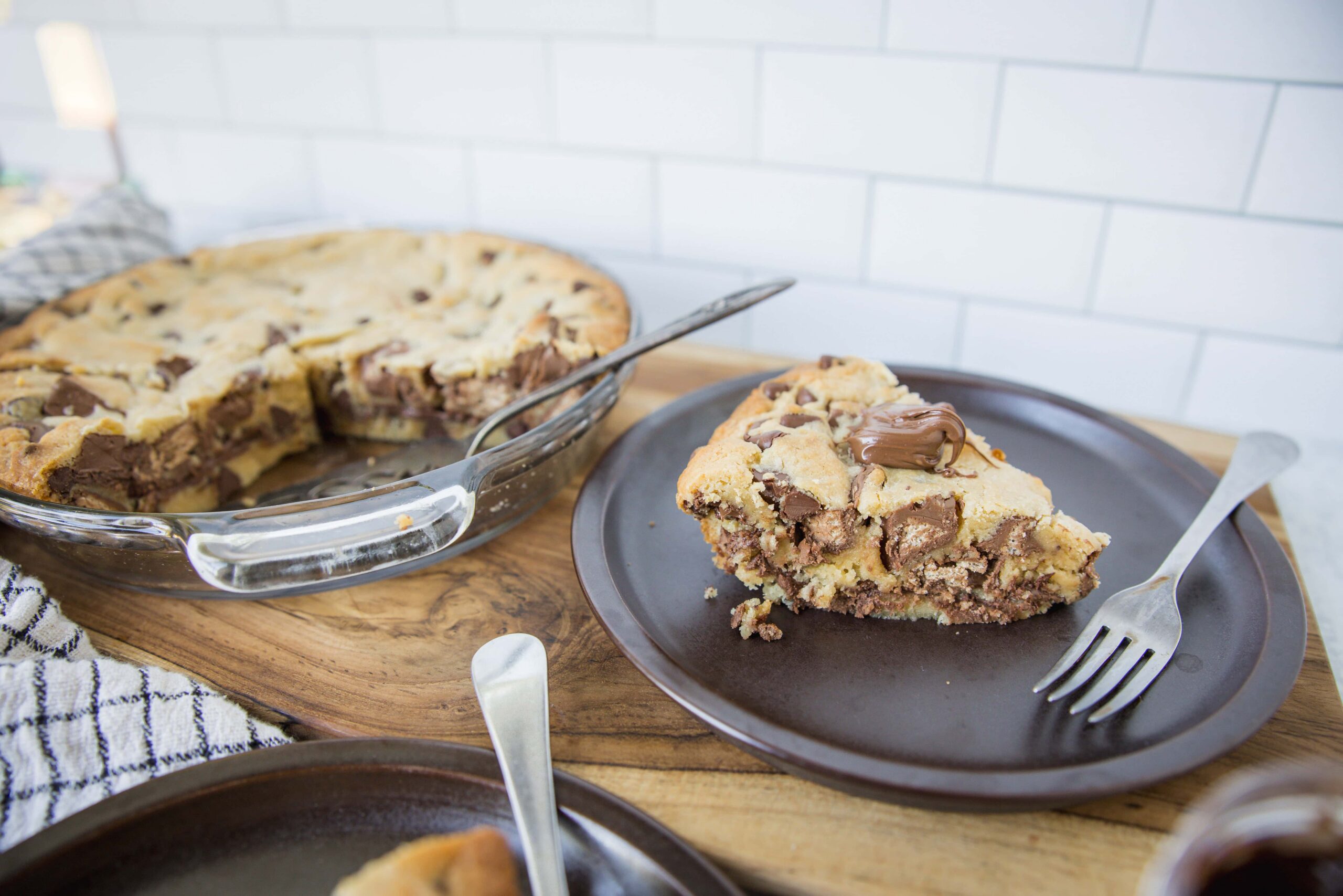 Chunks of homemade chocolate chip cookie dough cheesecake on a dark plate with a fork, with the full pie in a glass baking dish in the background, on a rustic wooden table with a white tiled wall.