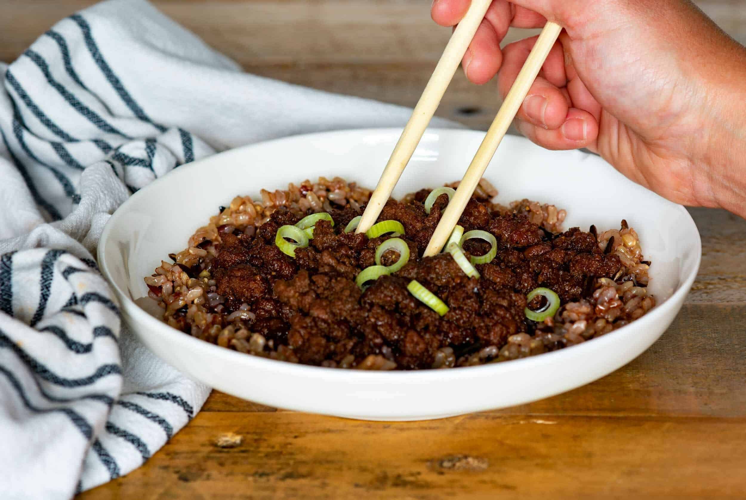 Savory ground beef and rice bowl garnished with sliced green onions, served in a white ceramic dish with chopsticks, on a rustic wooden table with a striped cloth.