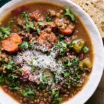 Hearty lentil vegetable soup with carrots, celery, and herbs served with rustic bread on wooden table. Perfect for comfort food or healthy meals, topped with grated cheese and fresh parsley.