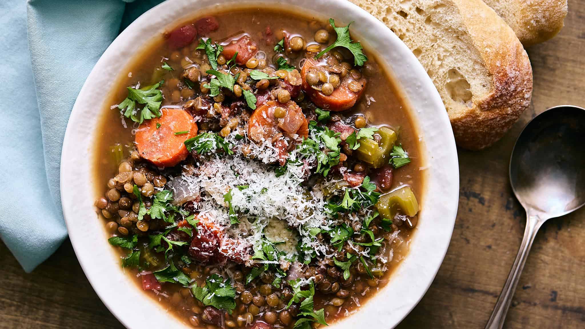 Hearty lentil vegetable soup with carrots, celery, and herbs served with rustic bread on wooden table. Perfect for comfort food or healthy meals, topped with grated cheese and fresh parsley.