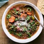 A bowl of hearty lentil vegetable soup garnished with fresh herbs, served with crusty bread on a rustic wooden table.