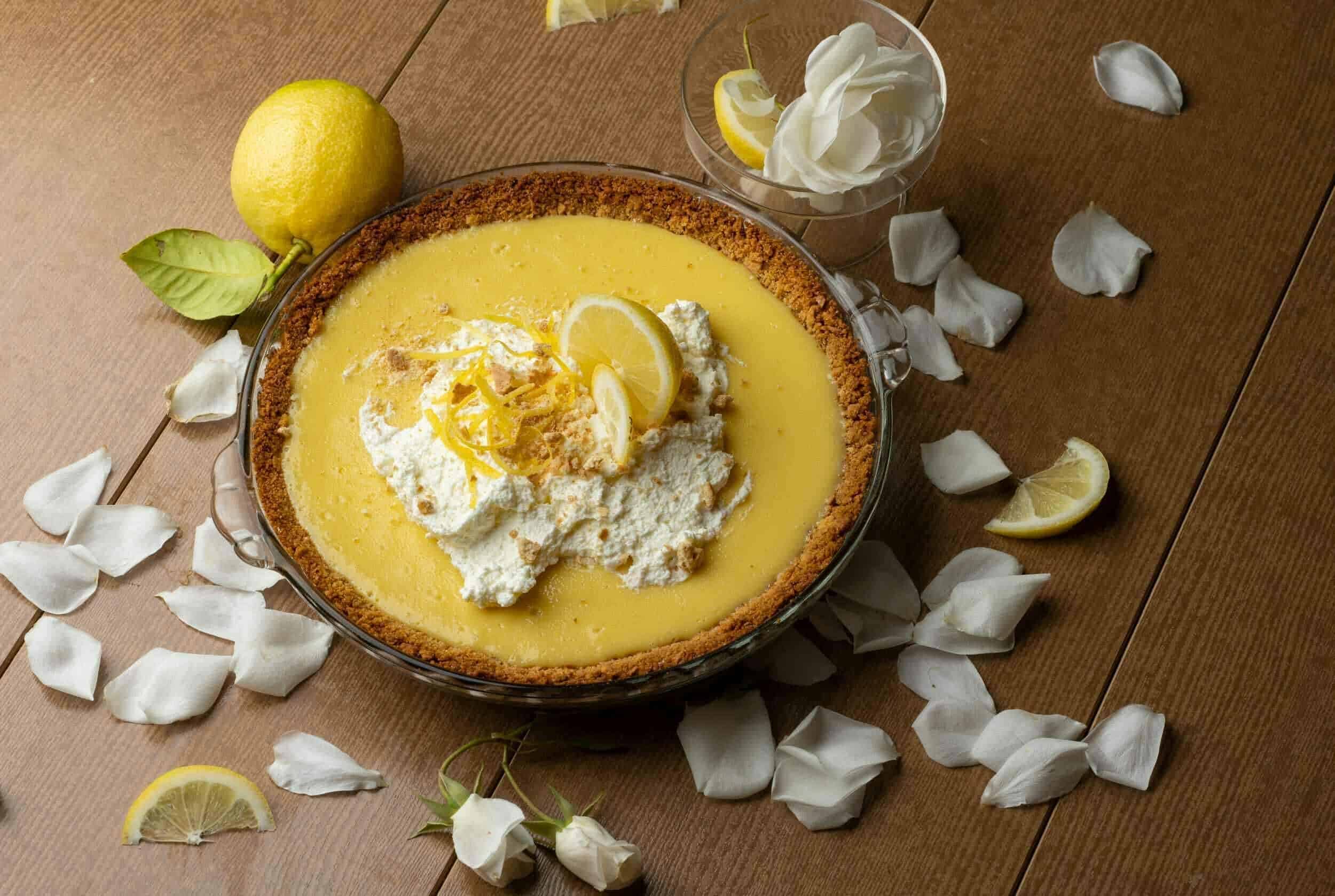 Lemon cheesecake with a graham cracker crust topped with whipped cream and lemon slices on a wooden table, surrounded by lemon wedges and white rose petals.