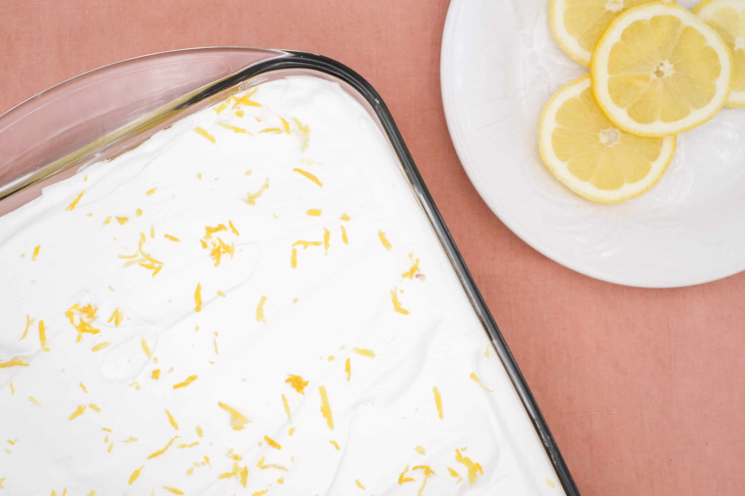 Freshly grated lemon zest on creamy dessert in a glass dish, with a plate of sliced lemons on the side, perfect for lemon-flavored recipes and dessert toppings.