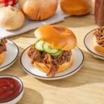 Tender shredded beef barbecue sandwiches on soft buns with fresh cucumber slices, served with ketchup, perfect for a classic bbq meal.