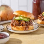 Tender shredded beef barbecue sandwich with fresh cucumber slices on a soft bun, served on a white plate with a side of ketchup and background vegetables for a delicious homemade meal.