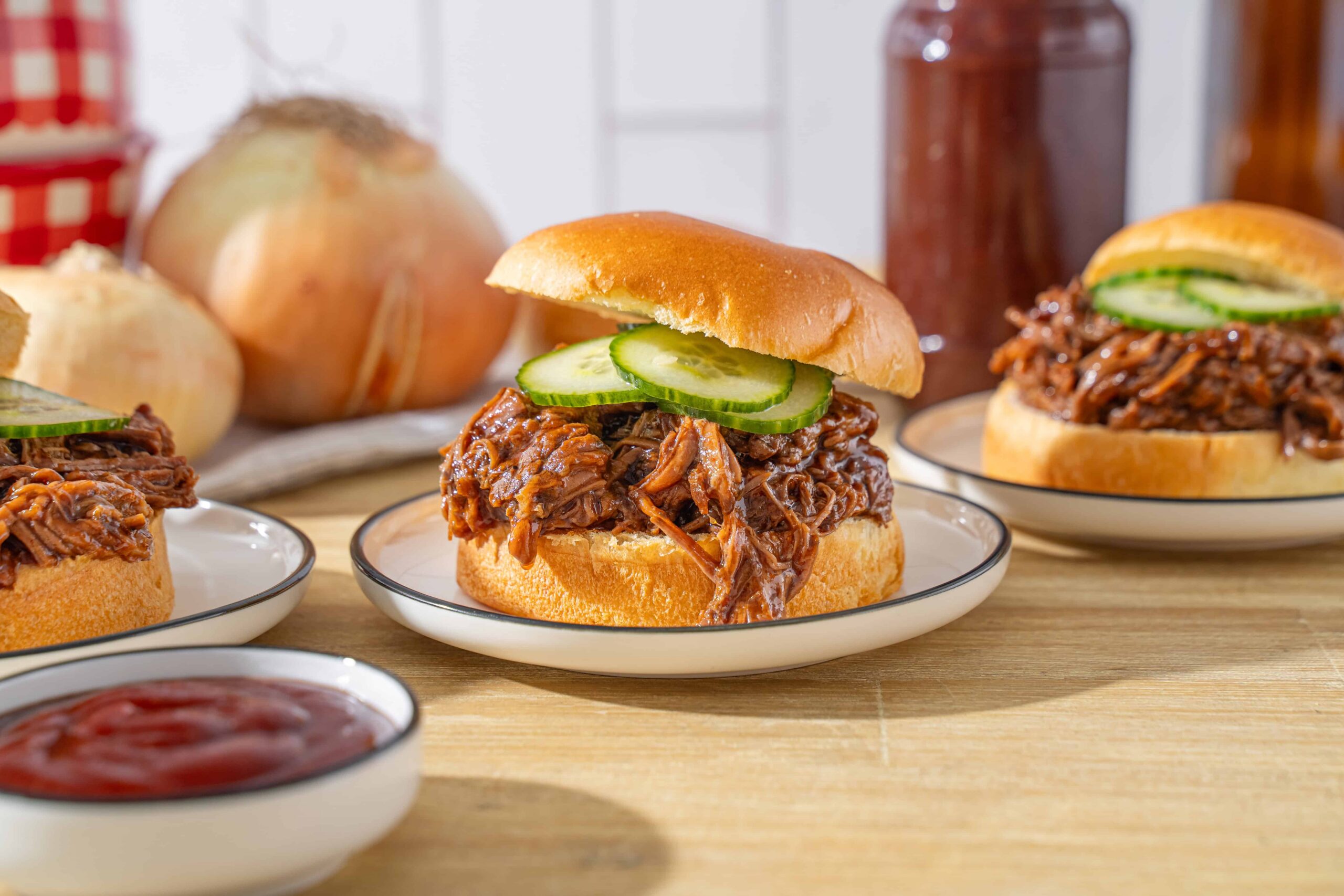 Tender shredded beef barbecue sandwich with fresh cucumber slices on a soft bun, served on a white plate with a side of ketchup and background vegetables for a delicious homemade meal.