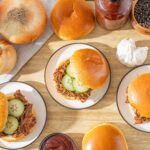 Slow-cooked shredded beef sandwiches with pickles on soft buns, surrounded by onions, garlic, black pepper, and tomato sauce on wooden table.