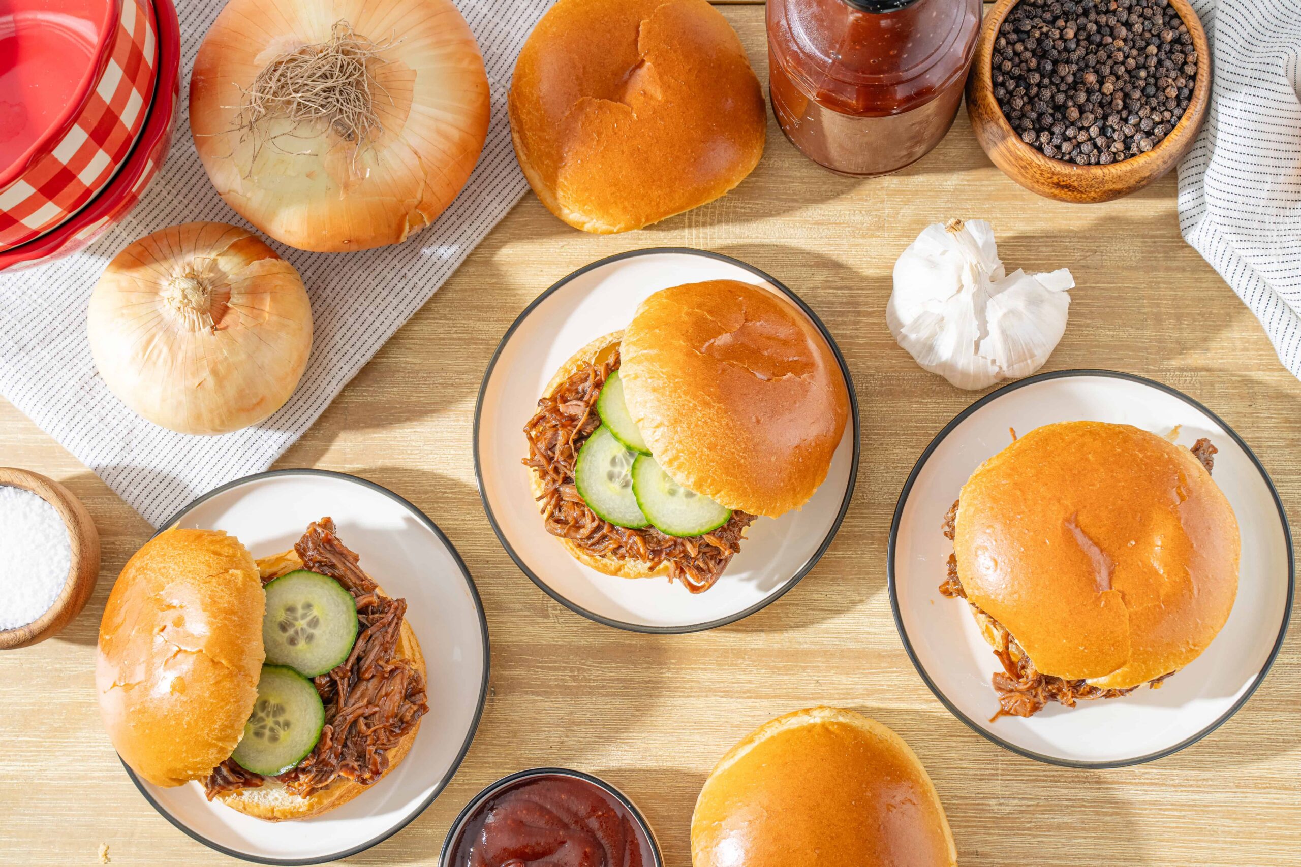 Slow-cooked shredded beef sandwiches with pickles on soft buns, surrounded by onions, garlic, black pepper, and tomato sauce on wooden table.
