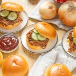 Slow cooker pulled pork sandwiches with cucumber slices on mini buns, served with barbecue sauce and fresh onions on a rustic table.