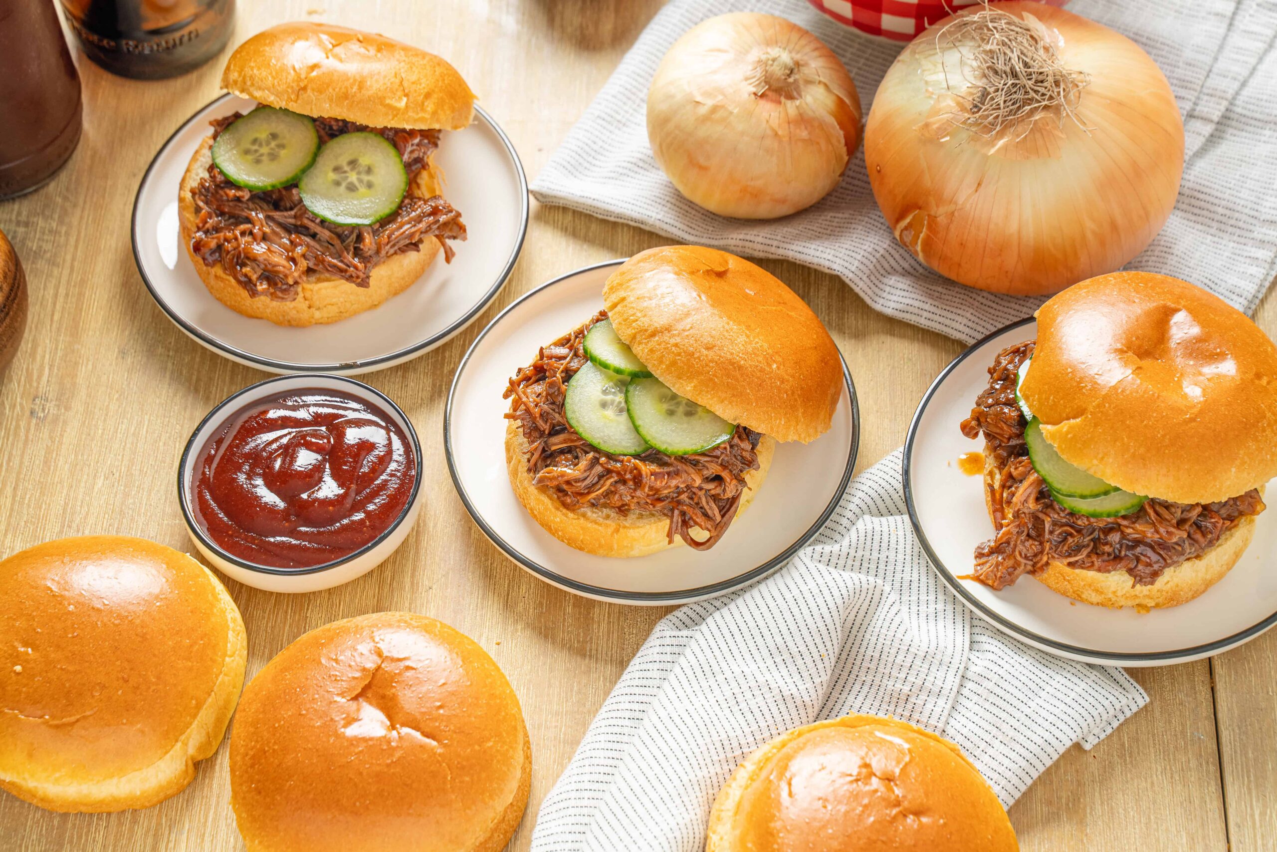 Slow cooker pulled pork sandwiches with cucumber slices on mini buns, served with barbecue sauce and fresh onions on a rustic table.