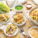 Creamy chicken and vegetable pasta casserole topped with cheese and crispy chicken bits, served in a large white baking dish with side plates of cooked pasta and shredded cheese.