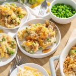 Creamy chicken and vegetable casserole with cheese, served in white bowls with green peas and shredded cheese toppings on a wooden table.