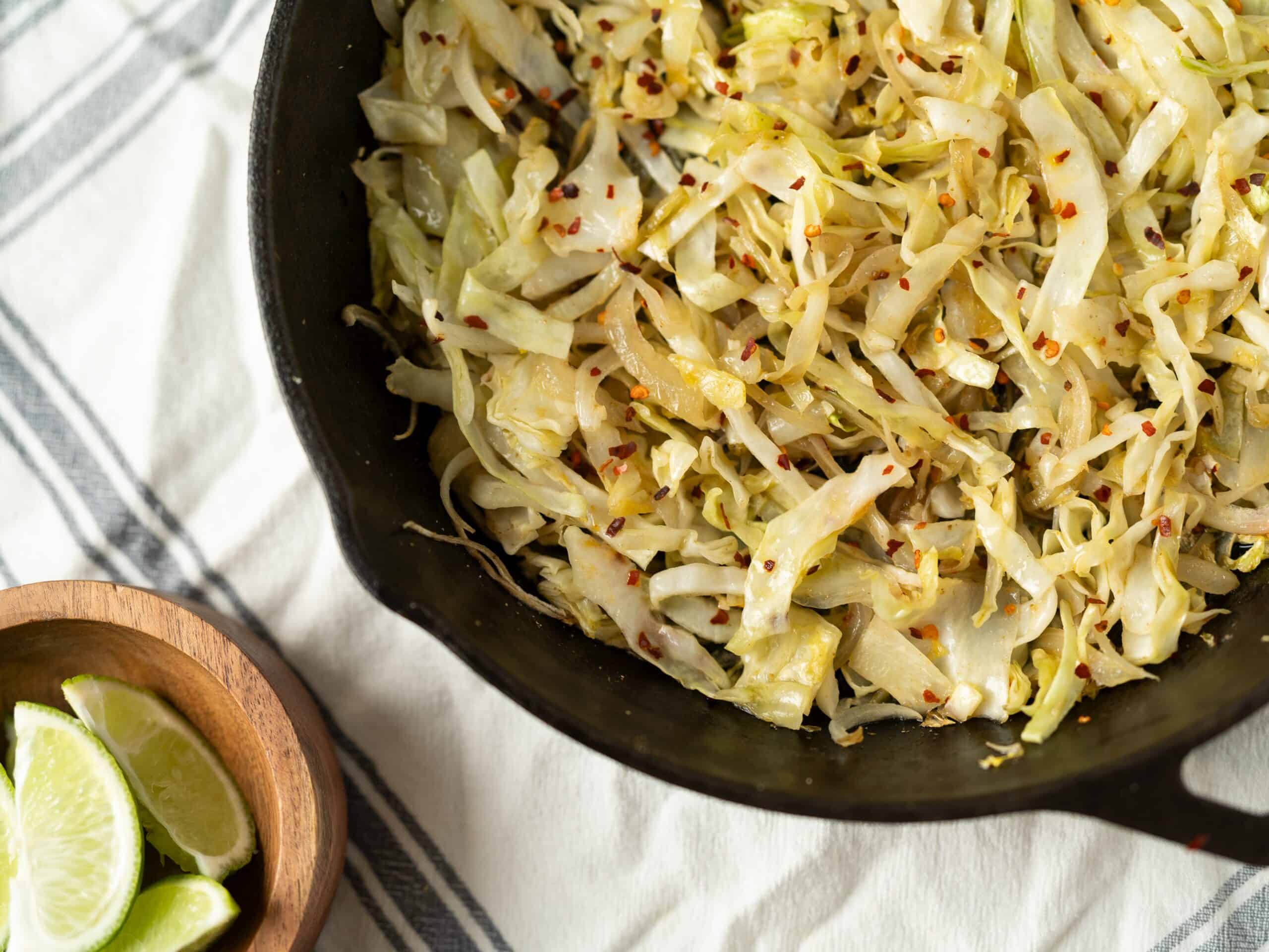 Sauteed cabbage with red pepper flakes in a black skillet, served with lime wedges on the side, perfect for healthy side dish or vegetarian meal.