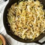 Sautéed cabbage with chili flakes in a black skillet on a striped cloth, garnished with lemon wedges on the side, making a flavorful and healthy vegetable side dish.