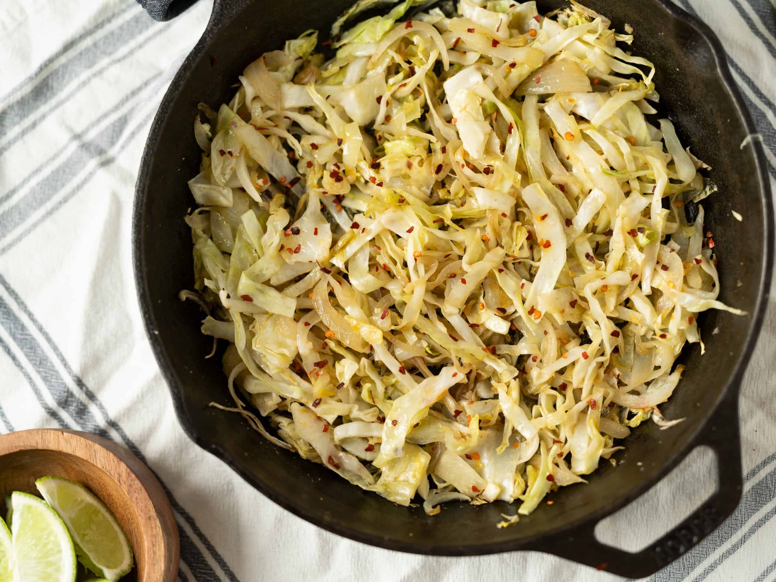 Sautéed cabbage with chili flakes in a black skillet on a striped cloth, garnished with lemon wedges on the side, making a flavorful and healthy vegetable side dish.