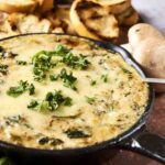 Creamy spinach and mushroom dip in a cast iron skillet garnished with fresh herbs, served with toasted bread slices and whole mushrooms on a rustic wooden board - perfect for appetizer or snack.