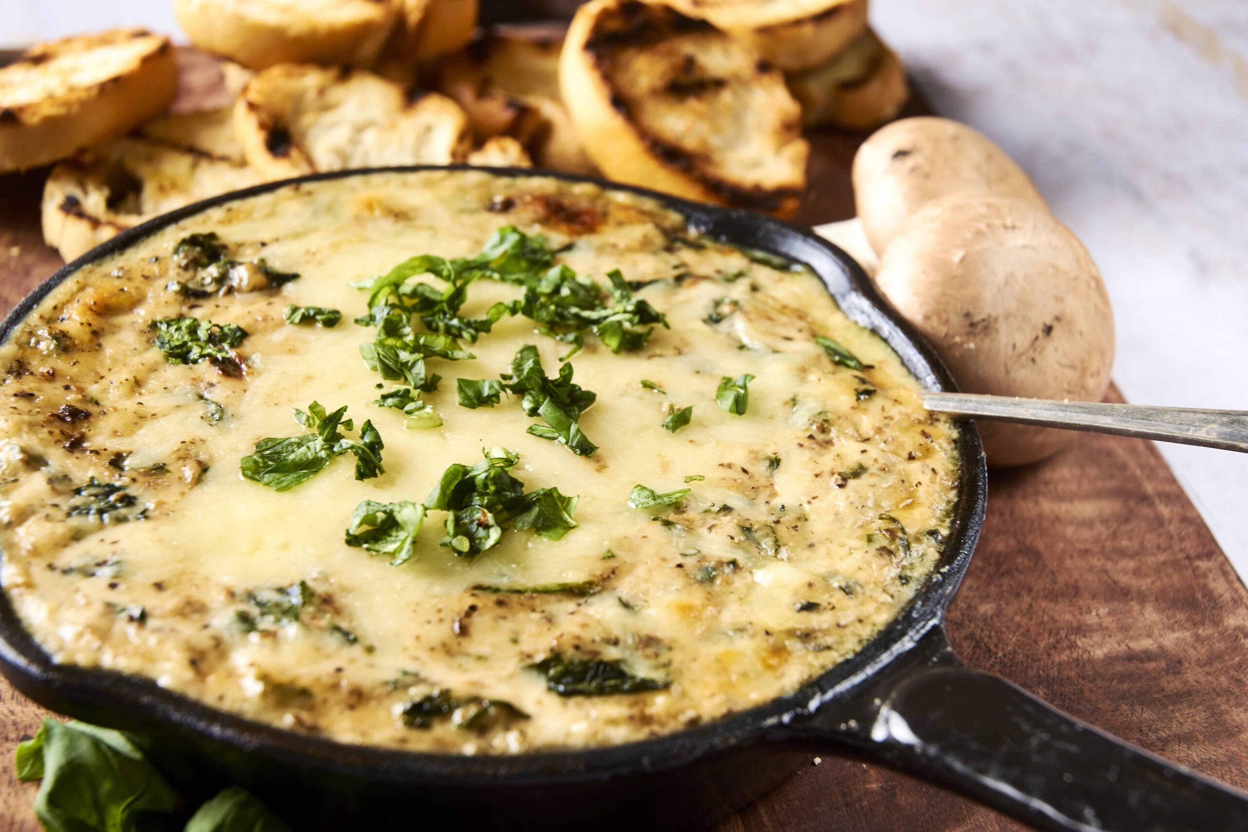 Creamy spinach and mushroom dip in a cast iron skillet garnished with fresh herbs, served with toasted bread slices and whole mushrooms on a rustic wooden board - perfect for appetizer or snack.