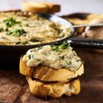 Creamy mushroom and herb topping served over toasted bread slices, with a cast iron skillet of mushroom evidence in background for hearty vegetarian appetizer or snack.