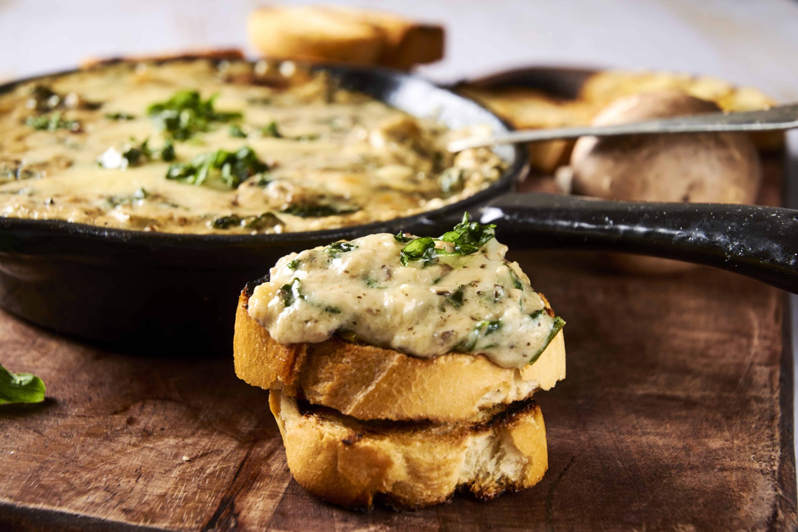 Creamy mushroom and herb topping served over toasted bread slices, with a cast iron skillet of mushroom evidence in background for hearty vegetarian appetizer or snack.
