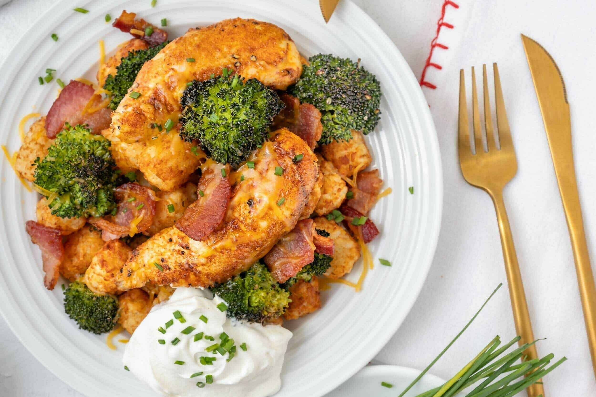 Grilled chicken with broccoli, bacon, and shredded cheese on a white plate, served with a dollop of sour cream, garnished with chives, and set on a white tablecloth with gold cutlery for a delicious meal.