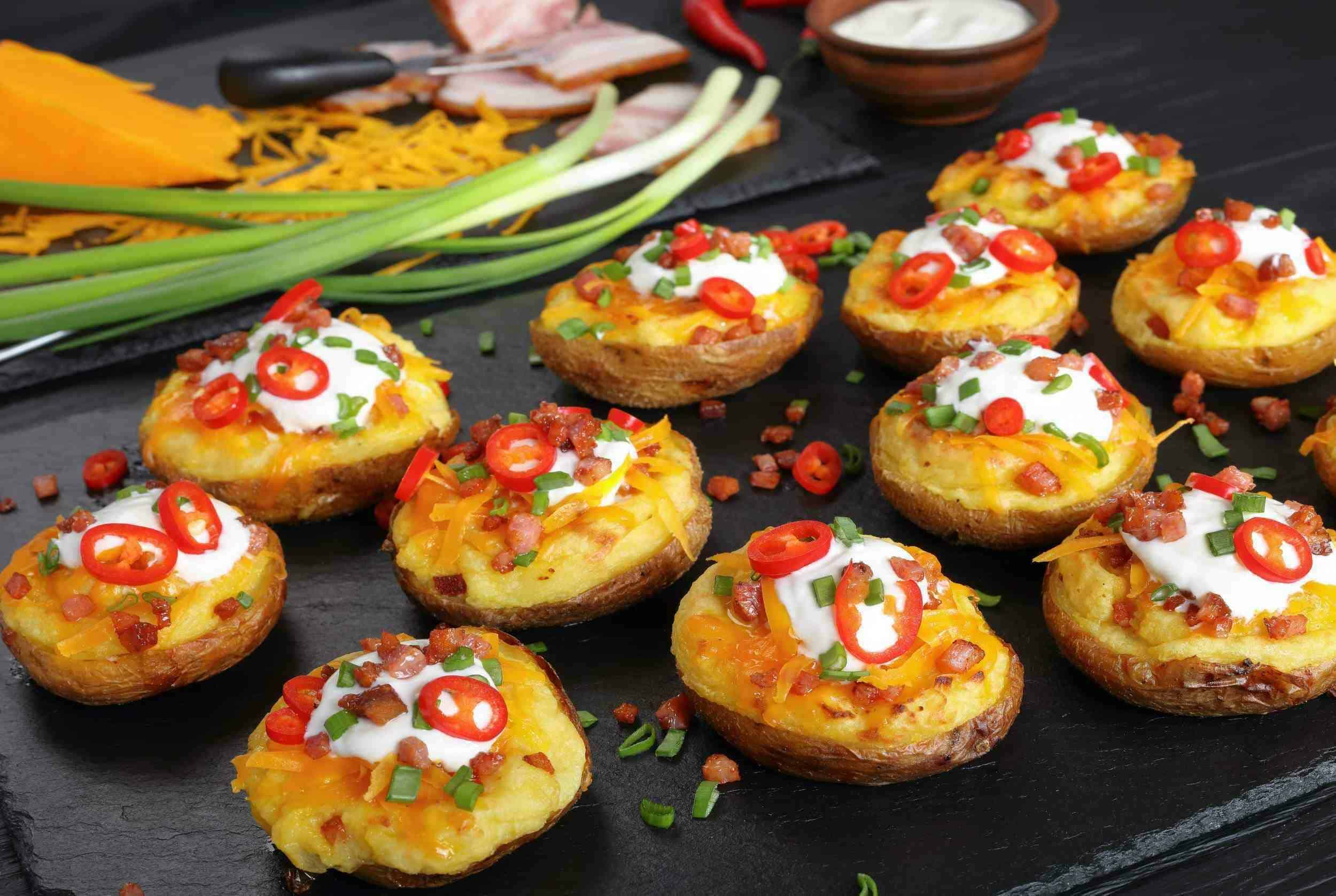 Delicious loaded mashed potato skins garnished with sour cream, chopped green onions, sliced red chilies, bacon bits, and shredded cheese on a black slate serving board.
