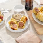 Sausage stuffed puff pastry rolls served with mustard dipping sauce on white plates, fresh and savory breakfast or snack, crispy golden pastry with juicy sausage filling, on a woven fabric tablecloth with a wooden cutting board, perfect for a brunch spread.