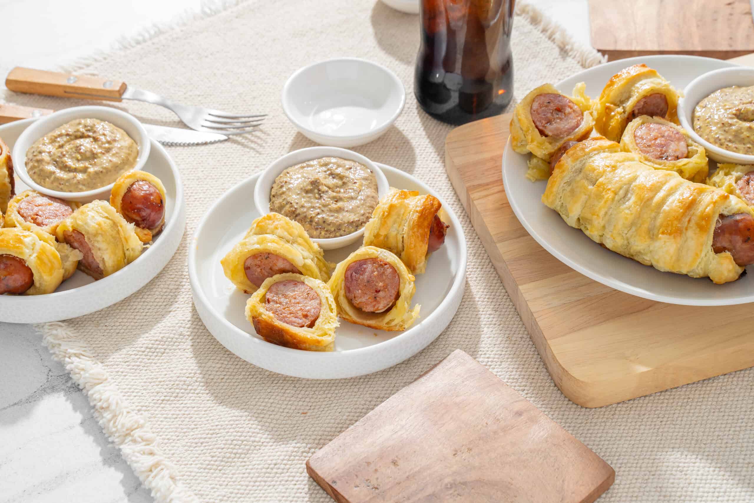 Sausage stuffed puff pastry rolls served with mustard dipping sauce on white plates, fresh and savory breakfast or snack, crispy golden pastry with juicy sausage filling, on a woven fabric tablecloth with a wooden cutting board, perfect for a brunch spread.