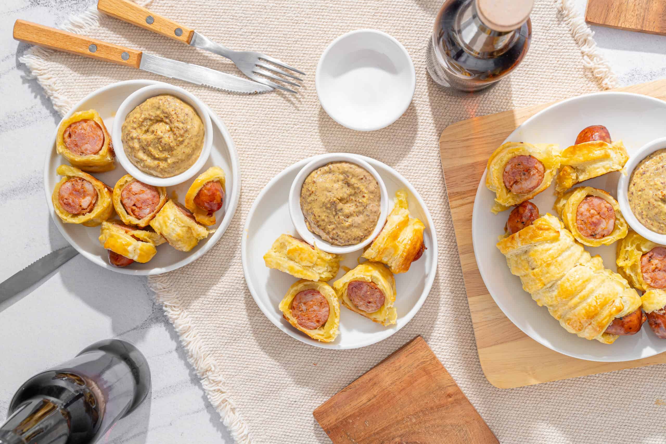 Air fryer sausage rolls with mustard sauce on white plates, served on wooden boards, with condiments and cutlery, ready for a tasty snack or appetizer.