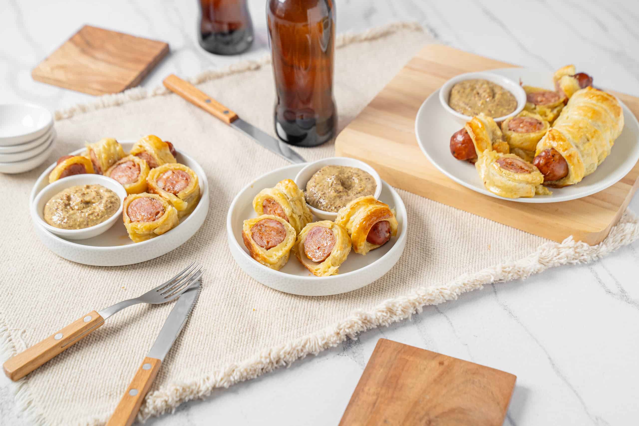 Savory sausage & cheese puff pastry bites served with mustard dipping sauce on white plates, garnished with wooden serving boards and neutral table setting for appetizer or party snack.