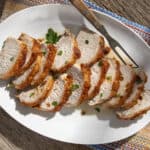 Juicy roasted pork tenderloin sliced on a white oval plate garnished with fresh parsley, served on rustic wooden table with colorful woven fabric for a delicious homemade meal.