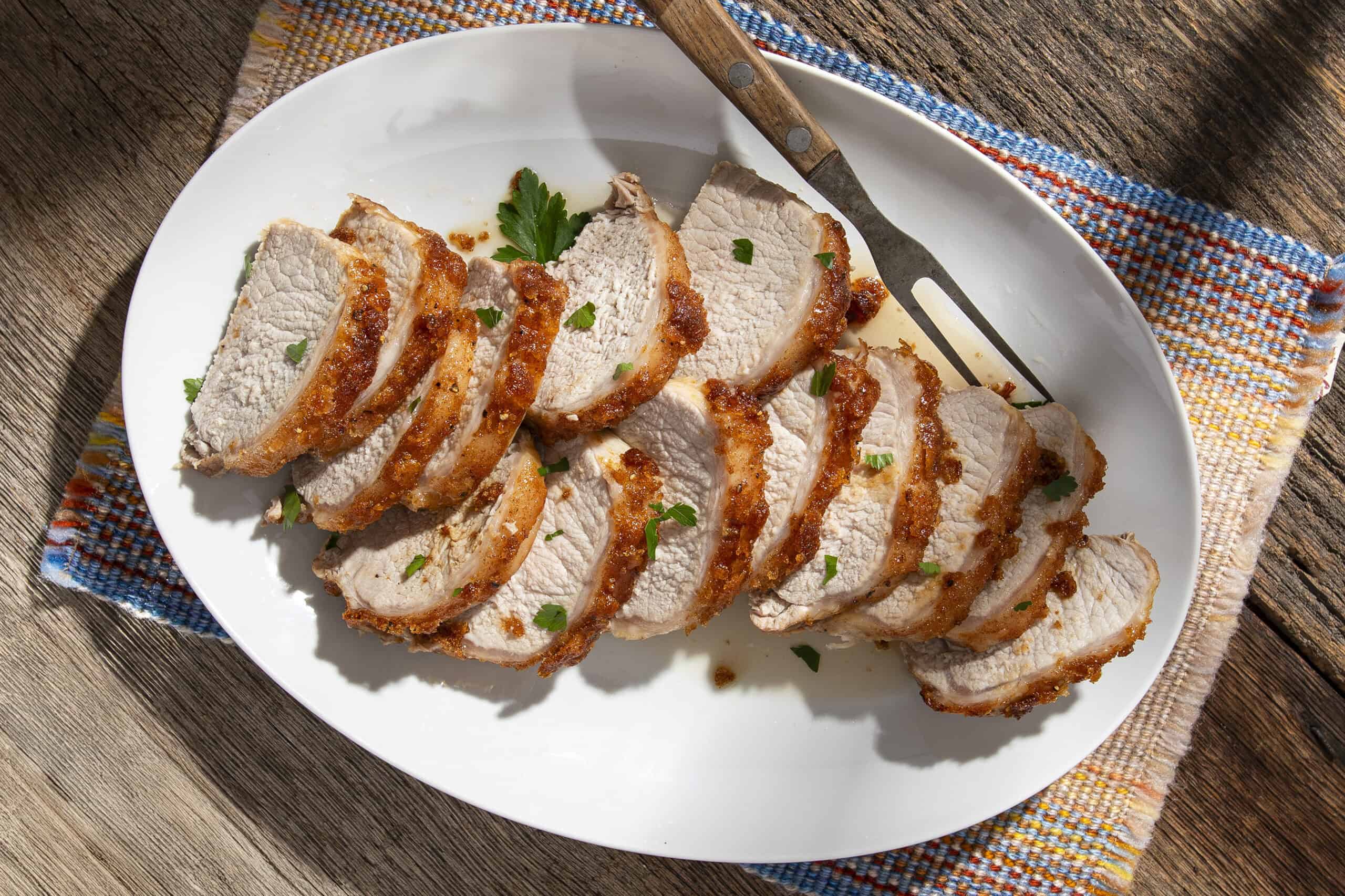 Juicy roasted pork tenderloin sliced on a white oval plate garnished with fresh parsley, served on rustic wooden table with colorful woven fabric for a delicious homemade meal.