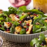 Fresh mixed green salad with feta cheese, pomegranate seeds, walnuts, and mixed greens in a white bowl on a woven placemat, with additional ingredients and drink in the background.