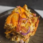 Golden baked casserole with melted cheese, crispy bacon bits, and red onion slices served on a textured black plate.