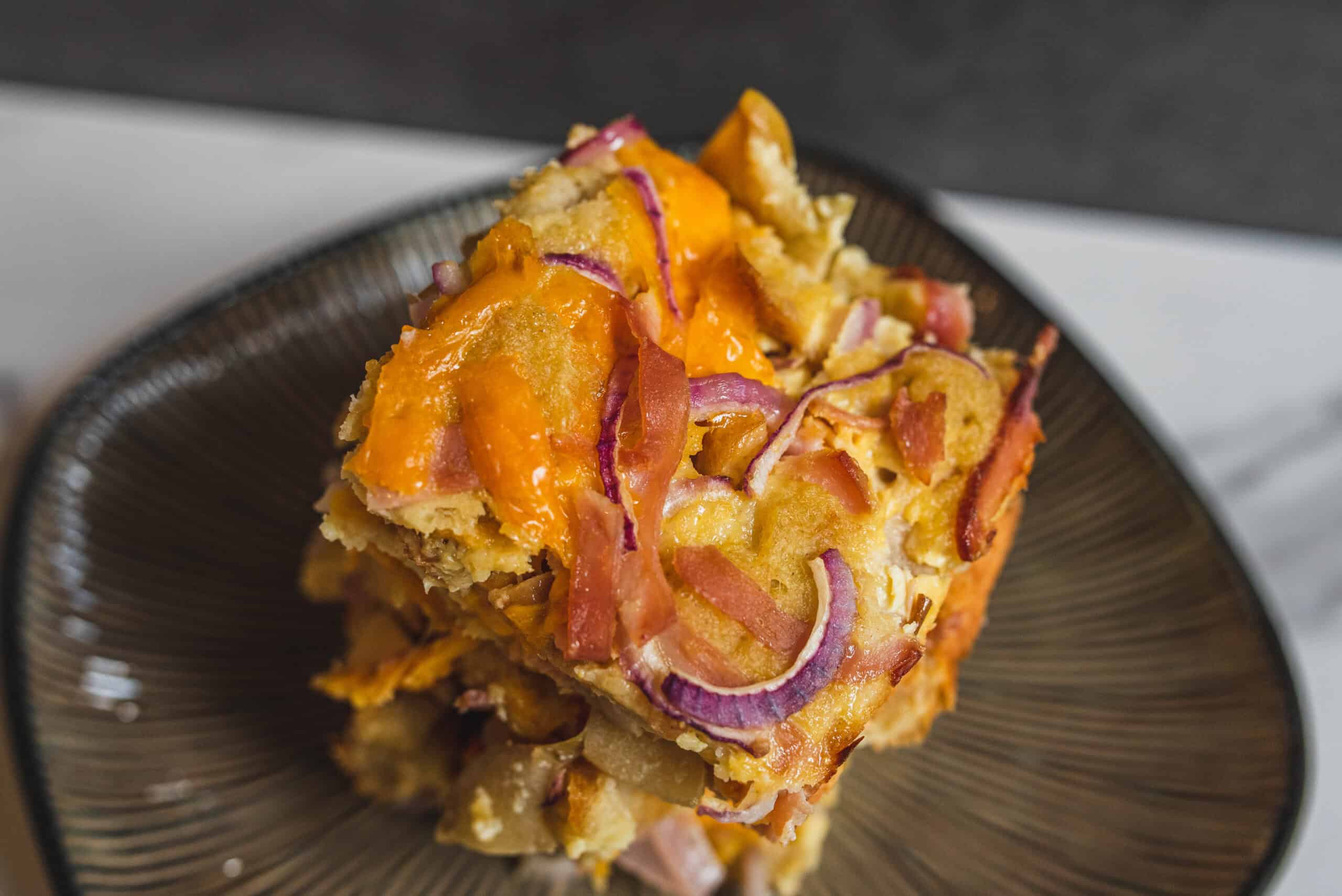 Golden baked casserole with melted cheese, crispy bacon bits, and red onion slices served on a textured black plate.