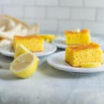 Soft lemon cake slices on white plates with lemon wedges, served on a light-colored table with fresh lemon slices and yellow-striped napkin, perfect for lemon desserts and baking recipes.