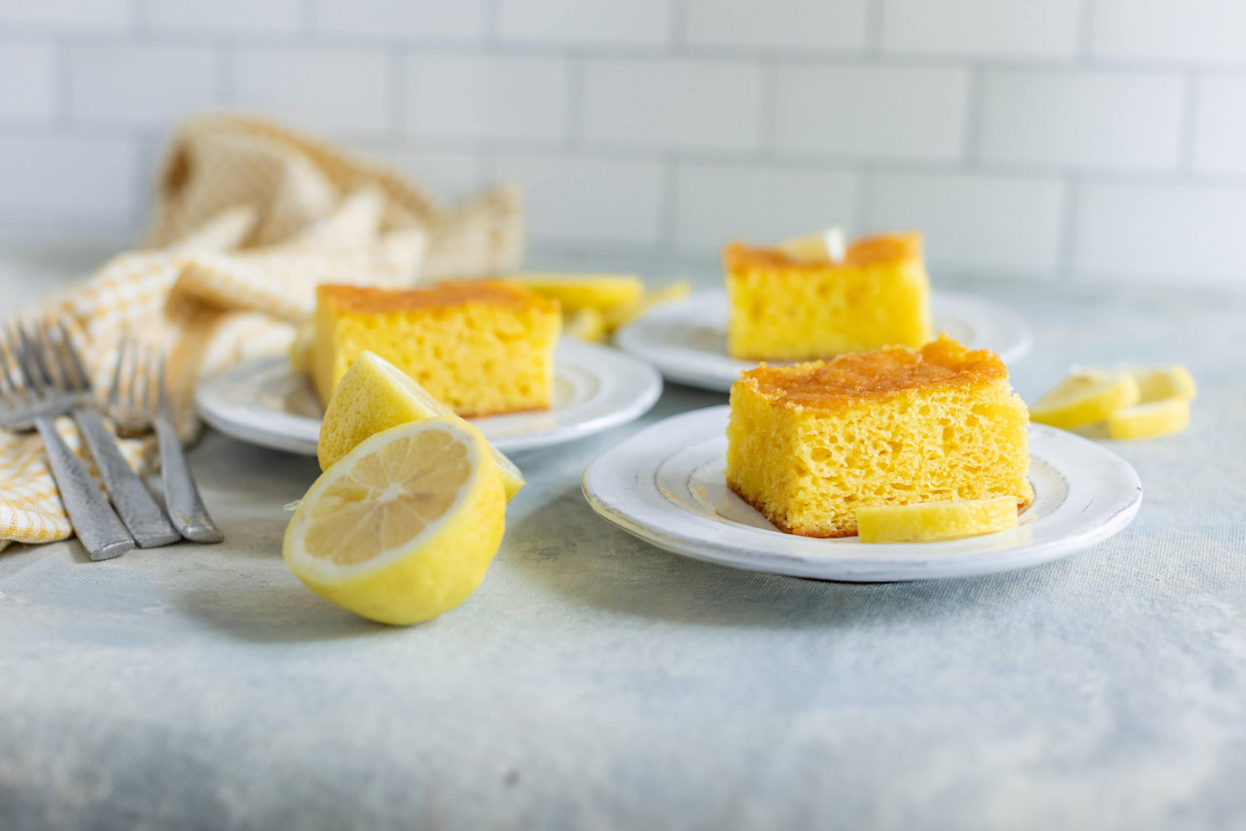 Soft lemon cake slices on white plates with lemon wedges, served on a light-colored table with fresh lemon slices and yellow-striped napkin, perfect for lemon desserts and baking recipes.