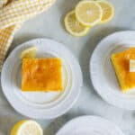 Lemon cake served on white plates with lemon wedges, fresh lemon slices, and a yellow checkered cloth for a bright, summery dessert presentation.