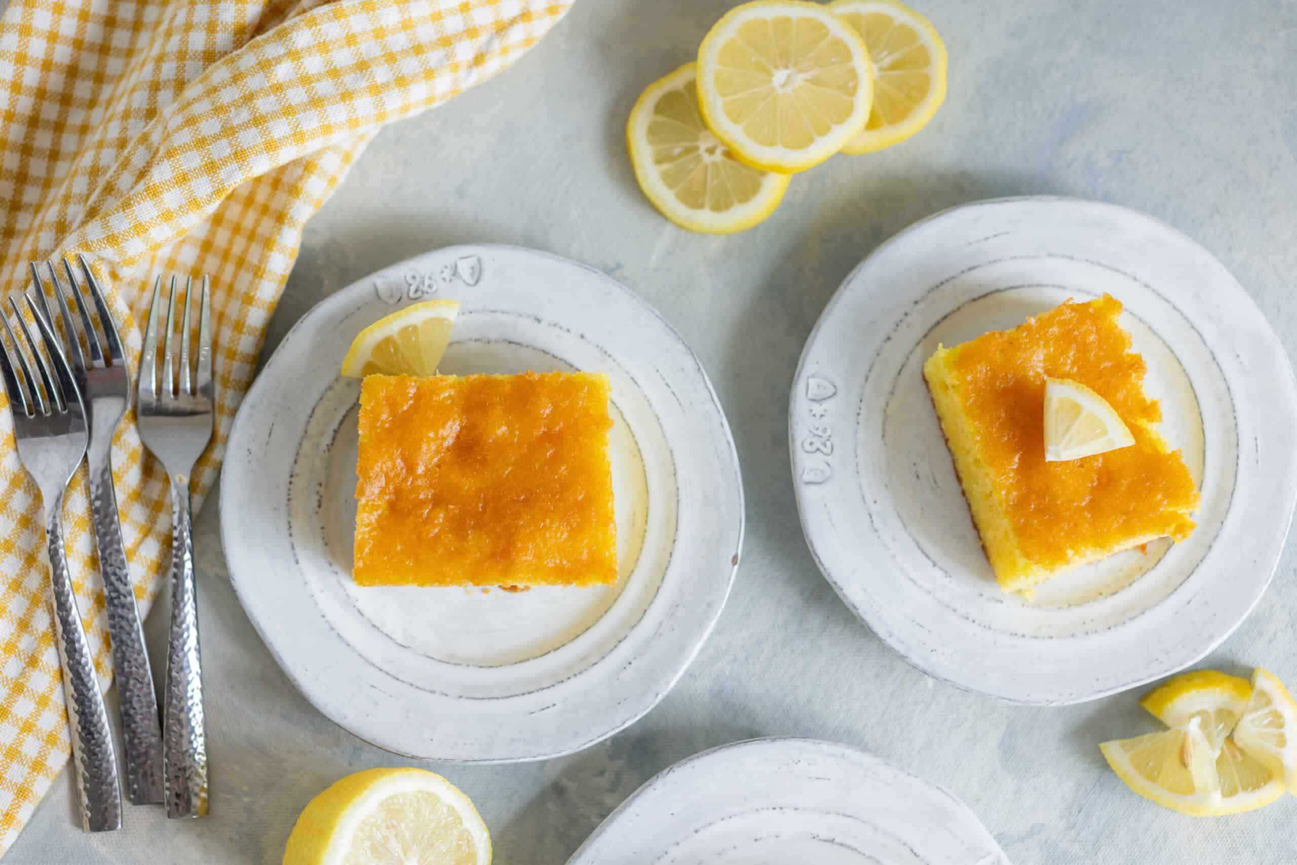 Lemon cake served on white plates with lemon wedges, fresh lemon slices, and a yellow checkered cloth for a bright, summery dessert presentation.